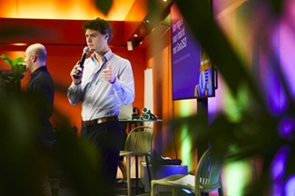   Spreker geeft een presentatie tijdens Esri Connect. De foto is genomen door bladeren heen, met kleurrijke verlichting in de ruimte.